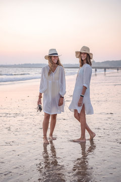 Graceful relaxed women in white dresses and hat smiling and looking at camera barefoot in water on beach in sunny day