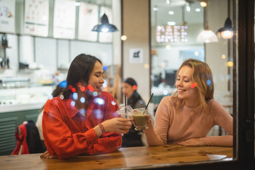 Young multiracial women smiling and speaking with each other while sitting at table in cozy cafe clinking cups and enjoying fruit drinks
