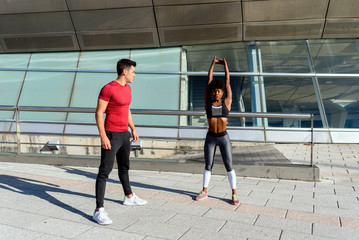 Young athletic man in red shirt giving support to active African American female in performing of stretching exercise during workout in sunny day in city