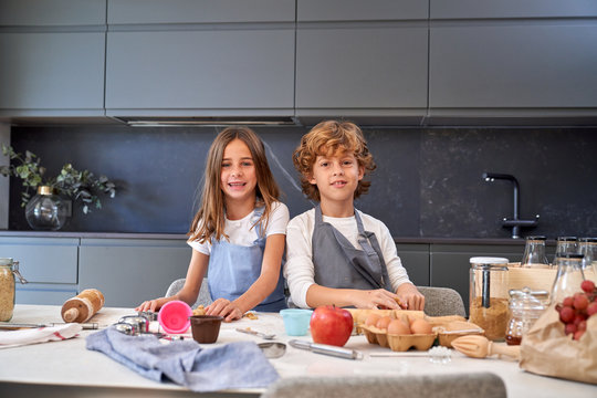 Little male and female kids in back splashes making paste while using different cooking implements at modern kitchen