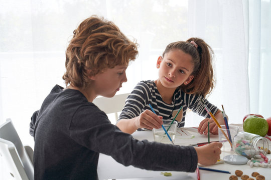Concentrated girl and boy in casual outfit painting with watercolor while sitting at table at home
