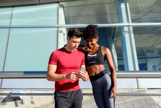 Personal trainer using smartphone and showing app to smiling fitness African American female while standing together next to building in sunny summer day