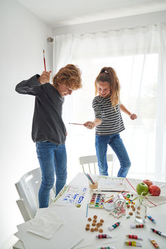 Delighted boy and girl in casual outfit looking at each other and crossing paintbrushes while standing on chairs near table with watercolor and playing at home