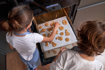 From above of faceless children in aprons spreading cookies on cookie sheet in oven at modern kitchen