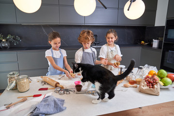 Shocked male and female kids in aprons looking at big cat on table with ingredients at modern kitchen