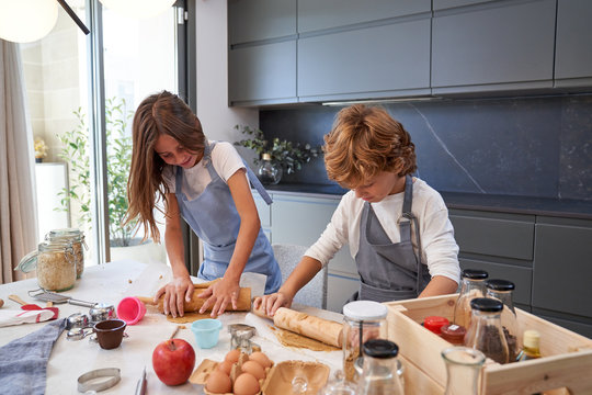 From above of male and female little kids in aprons smiling while rolling brown dough with pin on table at modern kitchen