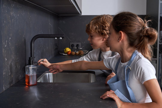 Side View Of Male And Female Kids In Aprons Preparing For Cook While Cleaning Hands In Sink At Modern Kitchen