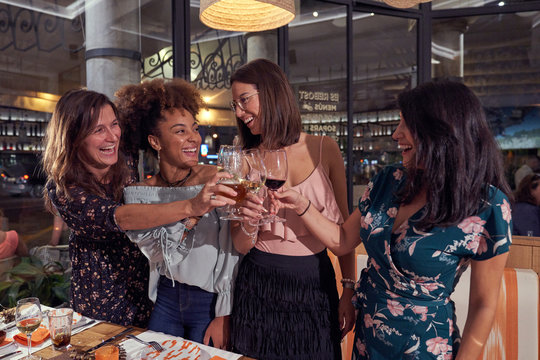 Multiethnic joyful female friends in casual clothing laughing while standing and proposing toast at stylish restaurant