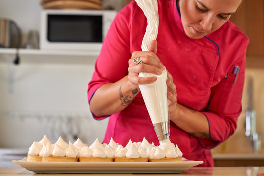 Woman chef in vibrant uniform using piping bag to filling tarts with whipped egg whites cream while cooking homemade delicious cookies with meringue in light modern kitchen