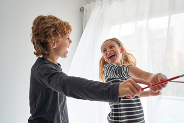 Excited boy and girl screaming and crossing paintbrushes while having fun near window at home