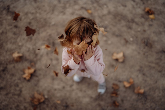 Active playful kid in pink warm clothes with closed eyes from pleasure throwing up autumn leaves in meadow in park