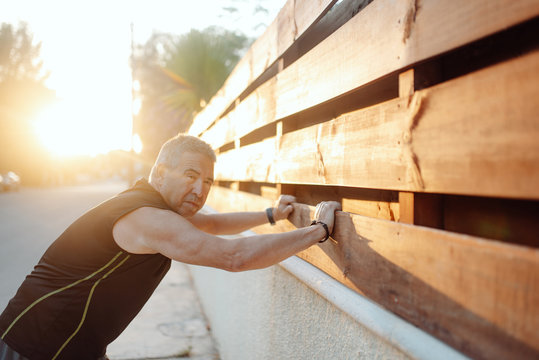 Enthusiastic active mature man in good sportive shape stretching in sunny street and wooden fence in summer day