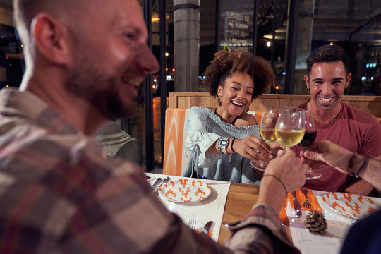 Multiethnic friends in casual clothing gathering at festive table and making toast at stylish restaurant