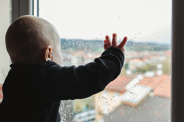 Unrecognizable baby in pajama touching wet glass and looking out window on rainy day at home