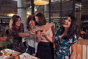 Multiethnic joyful female friends in casual clothing laughing while standing and proposing toast at stylish restaurant