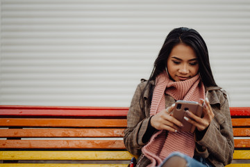 ethnic lady with suitcase sitting on rainbow bench and browsing smartphone on city street