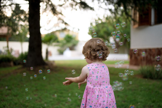 adorable back view child in pink dress capturing rainbow soap bubbles on green meadow in park
