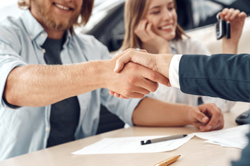 Obraz premium Car Salon. Young couple sitting at table man shaking hands with agent making agreement close-up blurred backgound smiling cheerful