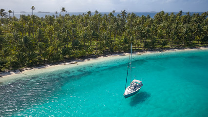 Sailing Yacht in San Blas Islands, Panama. A beautiful Aerial Shot of a white Sailboat anchored in a Blue Lagoon in front of a paradisiacal Tropical Caribbean Island overgrown with green Palm Trees. - obrazy, fototapety, plakaty