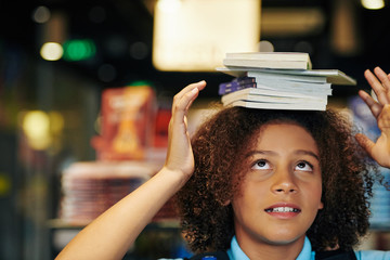 Horizontal portrait of cute teen girl with curly hair balancing stack of books on her head standing...