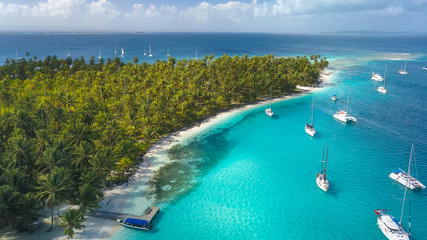 San Blas Islands, Panama - Drone Aerial View of many Sailboats & Sailing Yachts anchored in turquoise Water of Blue Lagoon next to white Sand Beach of Tropical Caribbean Island with green Palm Trees. - obrazy, fototapety, plakaty