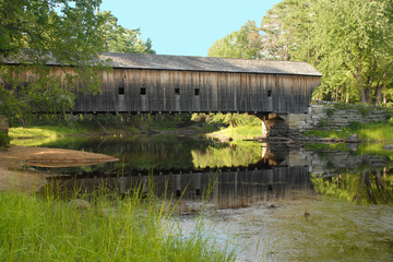 Old New England covered bridge over calm river. Reflection of bridge in water