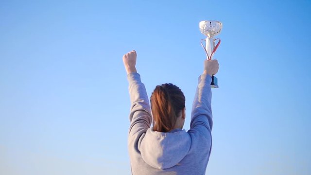 The girl runs and raises her hands with a golden cup over her head and celebrates her victory. Action in real time. The happiness of victory. The highest award. The joy of victory against a blue