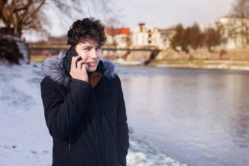 Teenage boy in winter near the river relaxing and talking on the phone, the guy smiles because he got the good news