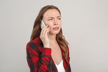 Freestyle. Young woman with freckles standing isolated on grey answering call on phone looking aside concerned