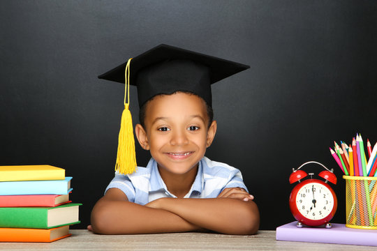 Young African American school boy sitting at desk with books, pencils and alarm clock on black background