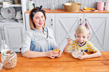 The boy greedily eats cereal with milk. Mother and son are smiling while having a breakfast in kitchen. Bright morning in the kitchen. Healthy Breakfast cereals and fresh fruit.