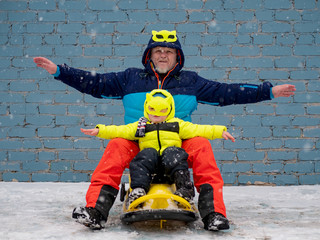 Dad and child in colorful winter clothes and mask riding sleigh along gray brick wall in snow. Man and boy waving hands depicting flight