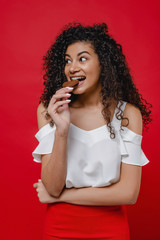 black woman biting heart shaped chocolate candy and smiling on red background