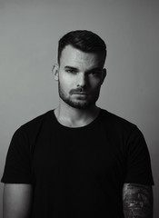 Black and white portrait of young and fit fashion model with tattoos on grey background. Portrait of pretty young guy with short hair - rembrandt lighting. Shot of man with grey tones