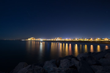 Fototapeta premium Night cityscape with sea and rocks in foreground and night star sky, night urban landscape of a Spanish city. Travel destination in Europe, card view with city lights and waterfront at night