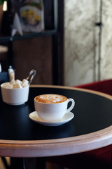 Cappuccino cup of coffee with sugar on the table in restaurant side view and a lot of copy space. Coffee cup latte and saucer. Espresso in cafe, dark background.