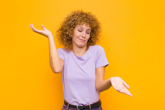 Young Afro Woman Shrugging With A Dumb, Crazy, Confused, Puzzled Expression, Feeling Annoyed And Clueless Against Orange Wall