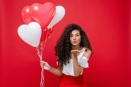 playful pretty black woman giving smooch kiss holding colorful helium balloons isolated on red