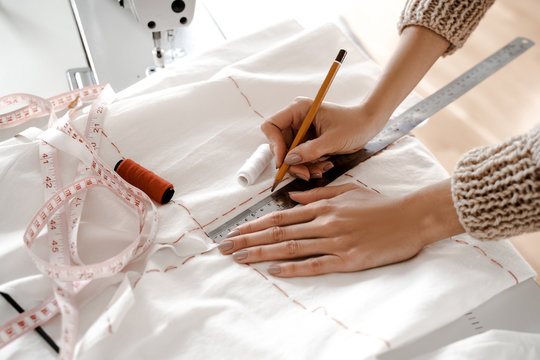 Closeup photo from above, hands on the seamstress table, creative space, hands draw markup for a new garment on a white cloth, make notes with a ruler and thread near the sewing machine