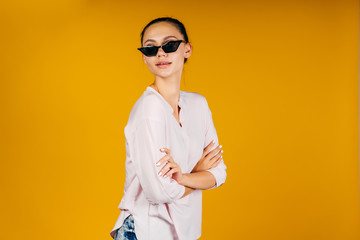 stylish girl in black glasses and a white blouse, clasping herself with his hands, looks at the camera. yellow background