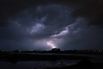 Naklejka na ściany i meble Thunderstorm with bright lightning striking behind a tree near a river