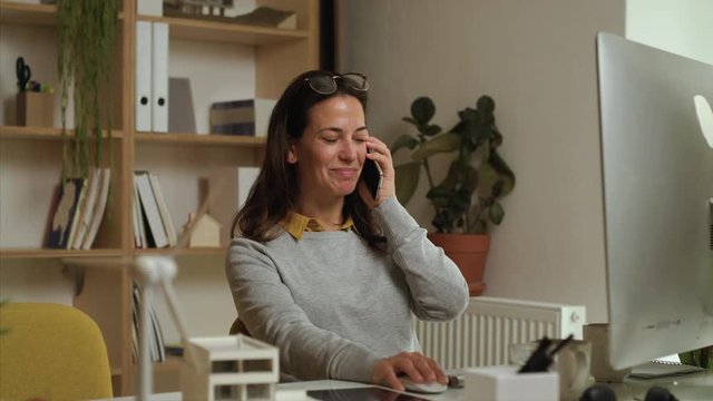 Attractive businesswoman sitting at the desk indoors in office, using smartphone.