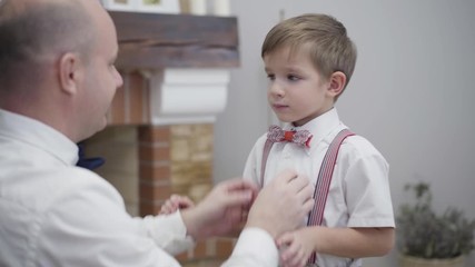 Close-up of confident Caucasian man helping little boy to tie a bow tie. Young father helping son to get ready for birthday party. Care, unity, support.