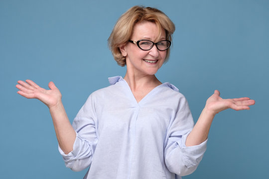 Senior mature caucasian woman in glasses and casual clothes shrugging her shoulders. Have no idea what to do in this situation. Studio shot