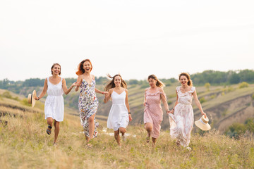 The company of cheerful female friends have a great time together on a picnic in a picturesque place overlooking the green hills. Girls in white dresses dancing in the field
