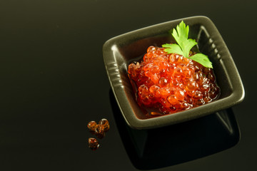 Fresh red caviar with a leaf of parsley in little black bowl on the black reflective background.