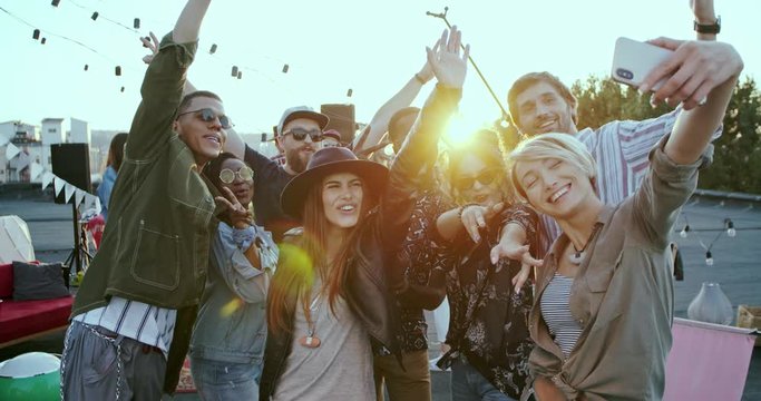 Cheerful young multiethnic people having fun and laughing while posing to the smartphone camera and taking selfie photos at the rooftop party.