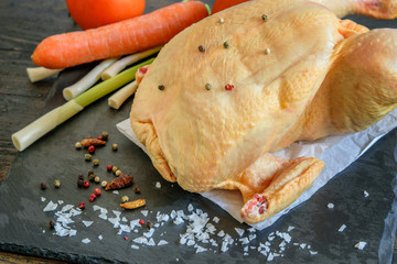 Whole uncooked chicken on the kitchen black board with vegetables tomatoes, carrot, fresh green garlic and spices closeup.