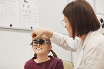 Portrait of female optometrist setting up trial frame while checking eyesight of little girl in...