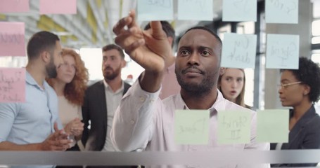 Male affrican team member thinking and replacing sticky notes with colleagues behind him. Group of diverse office staffs making project strategy at office meeting using glass board.